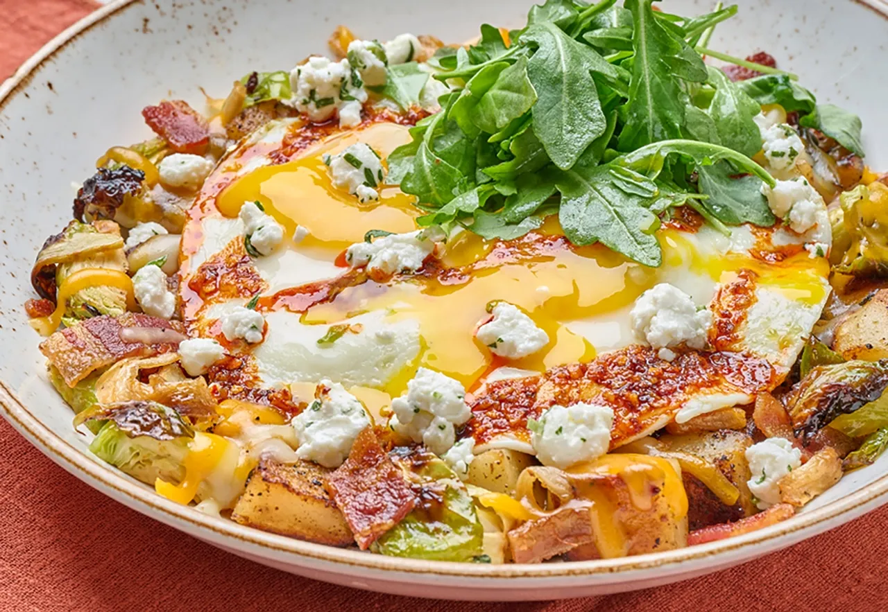 An aerial view of the First Watch Honey Bacon Brussels Hash topped with eggs and a drizzle of chili crisp, highlighting the deep, savory umami restaurant menu strategy